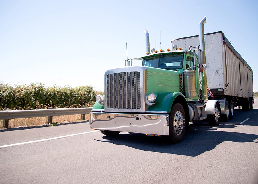 Classic American bonneted large green rig semi truck with high stylish chrome exhaust pipes transporting commercial cargo in bulk container trailer on the highway with trees behind the safety fence